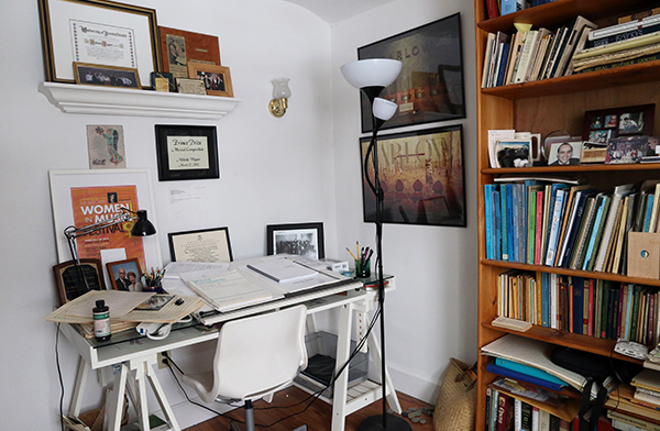 Photo of a desk with piles of manuscripts, a lamp, and a bookcase.