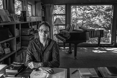 Aaron Holloway-Nahum sitting at a desk with Copland materials in a room with a bookcase, grand piano, and big window from which trees are visible.