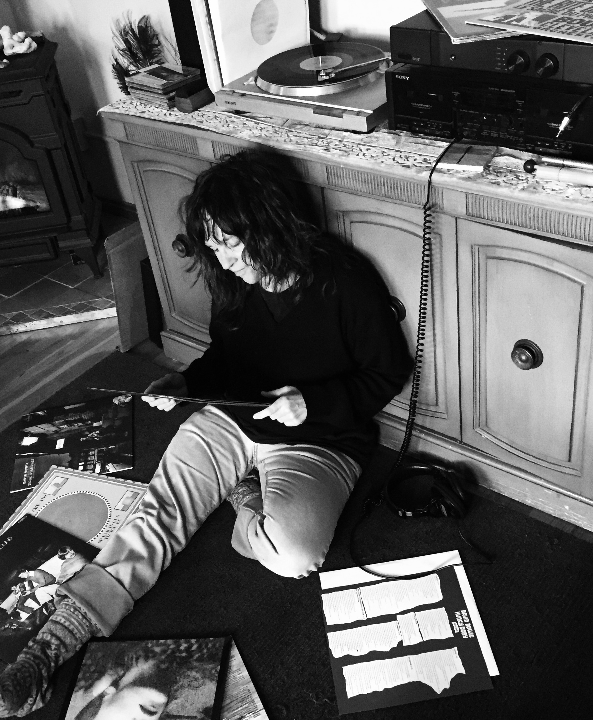 A black and photo photo of Gretta Harley sitting on the floor examining a pile of LPs; a turntable with an LP on it is in back of her.