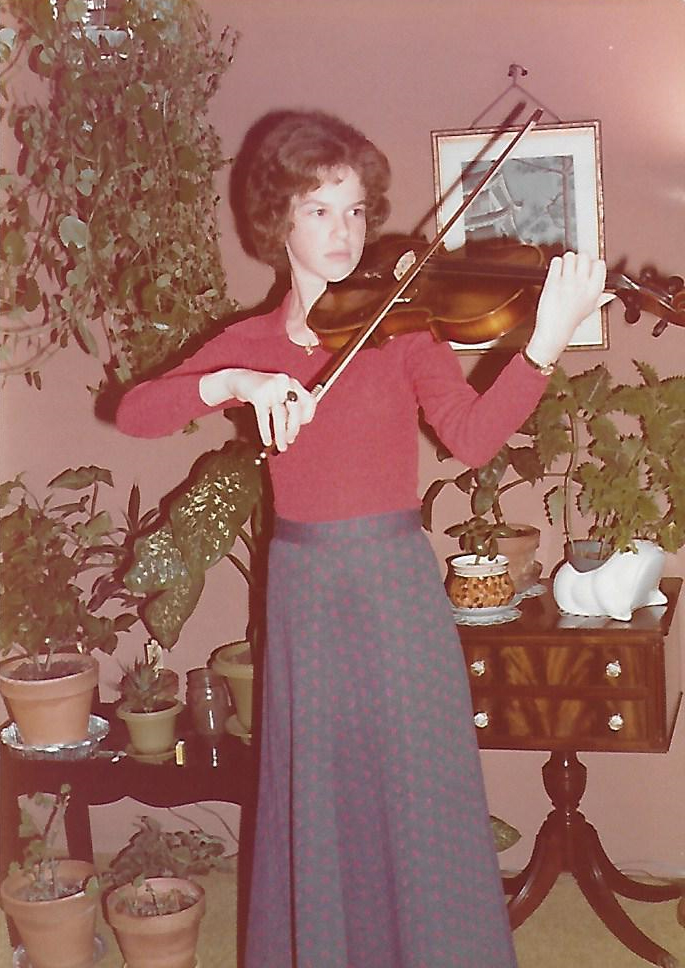 A very young Martha Mooke playing viola at home in front of a bunch of potted plants.
