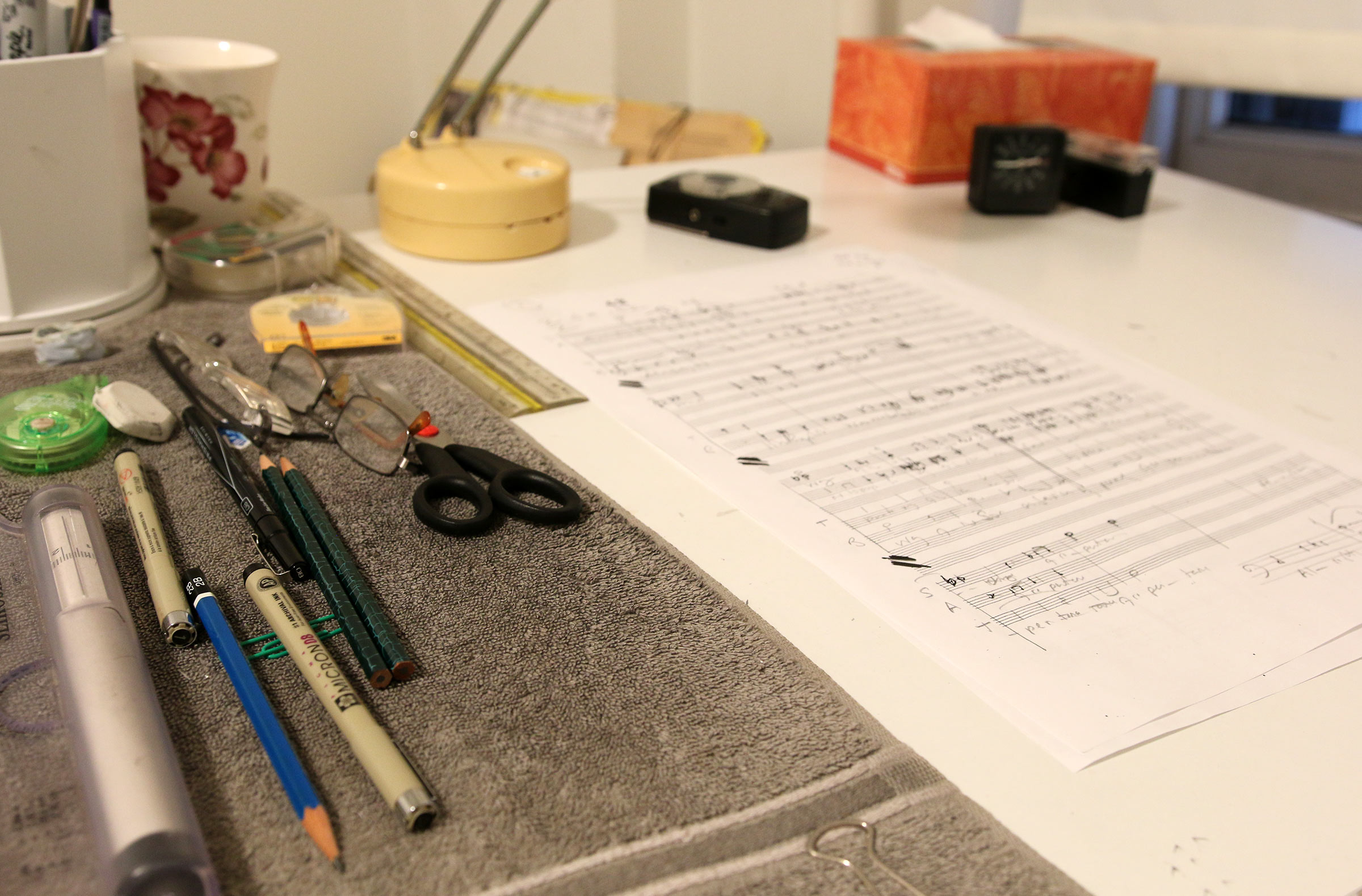 Pencils, a pair of glasses, scissors, a box of tissues and a sheet of music manuscript paper on a desk.