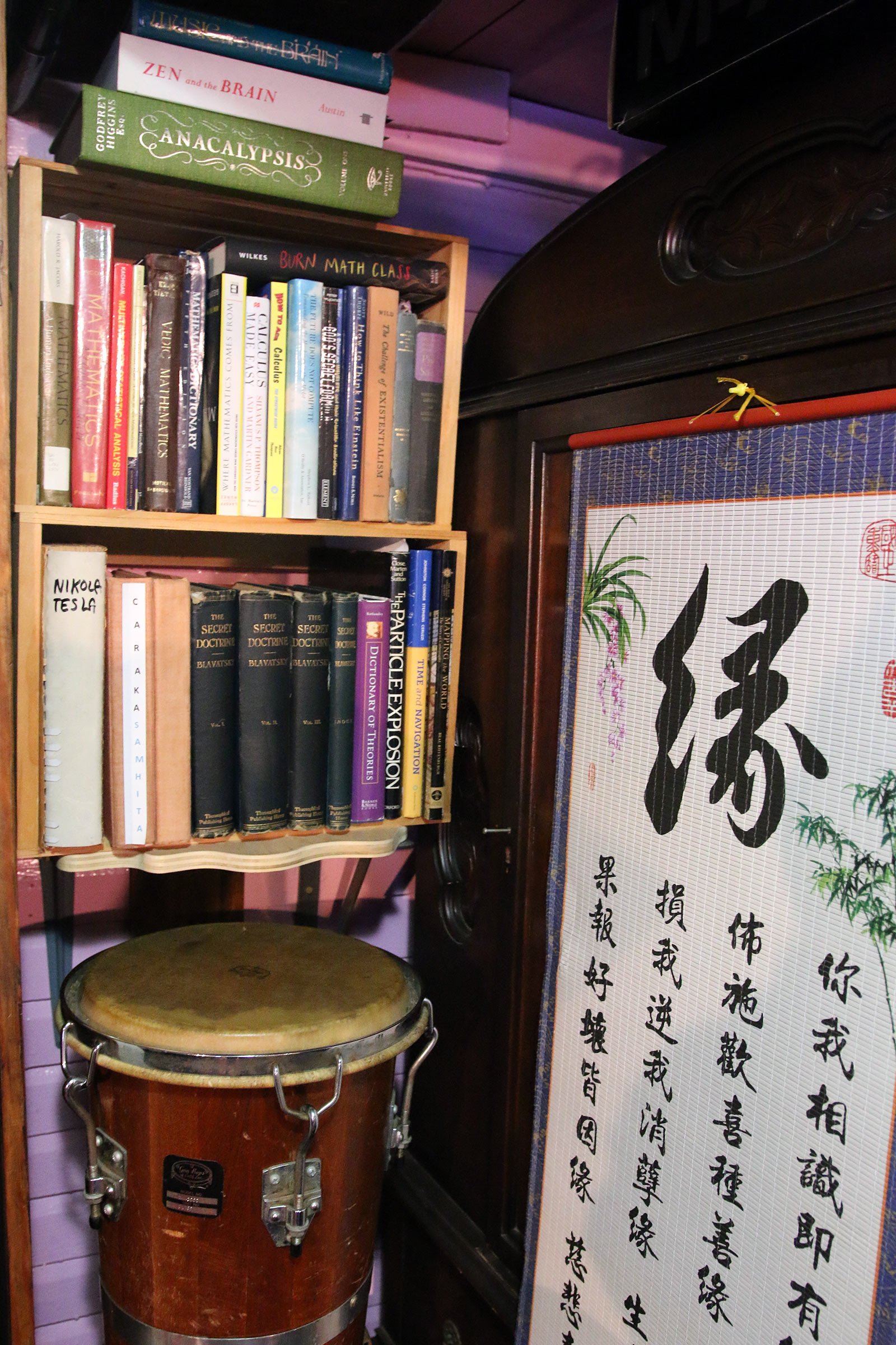 One of Milford Graves's drums on a shelf in a bookcase underneath two rows of books which is next to a Japanese scroll.