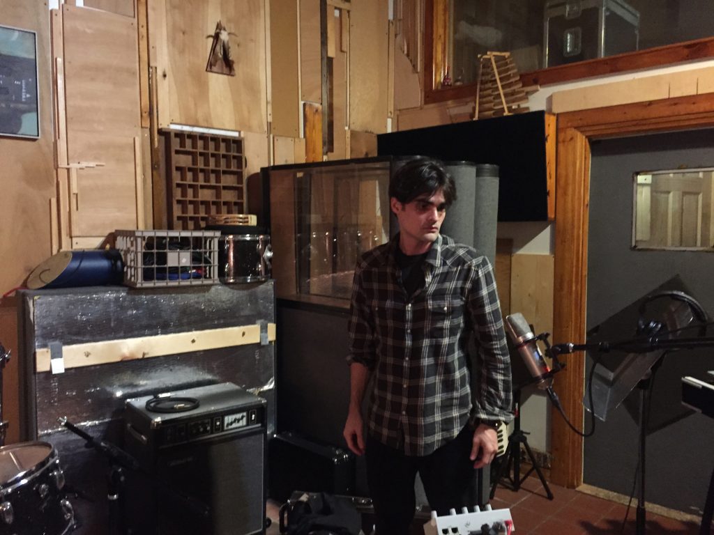 Noah standing in the middle of a recording studio which is cluttered with a drum, a music stand, a speaker and other pieces of electronic equipment. 