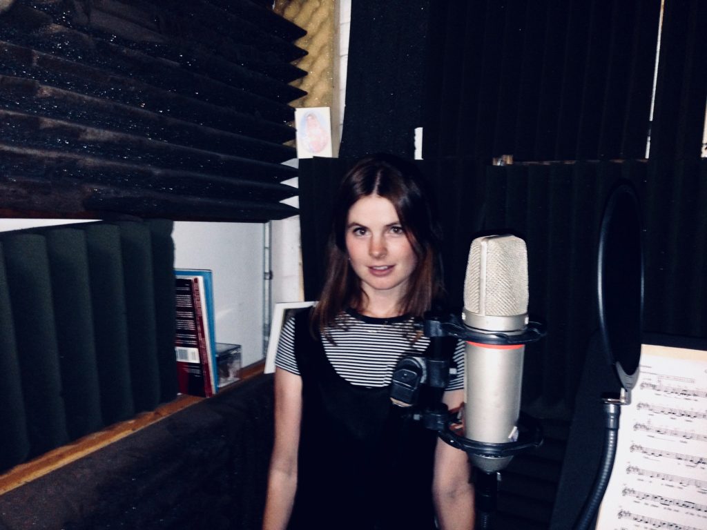 Rosie K standing in front of a microphone in a recording studio, which some sheet music nearby.