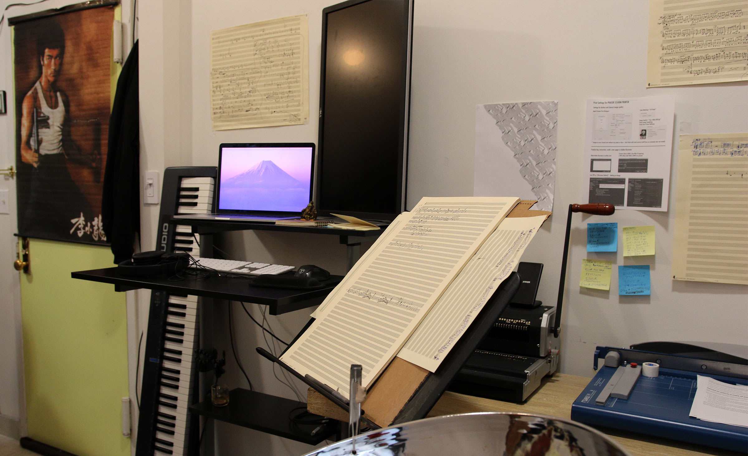 A view of the "office" part of Andy Akiho's apartment which includes a posted of Bruce Lee, a MIDI keyboard on its side, a computer terminal, some music stands, and handwritten scores.