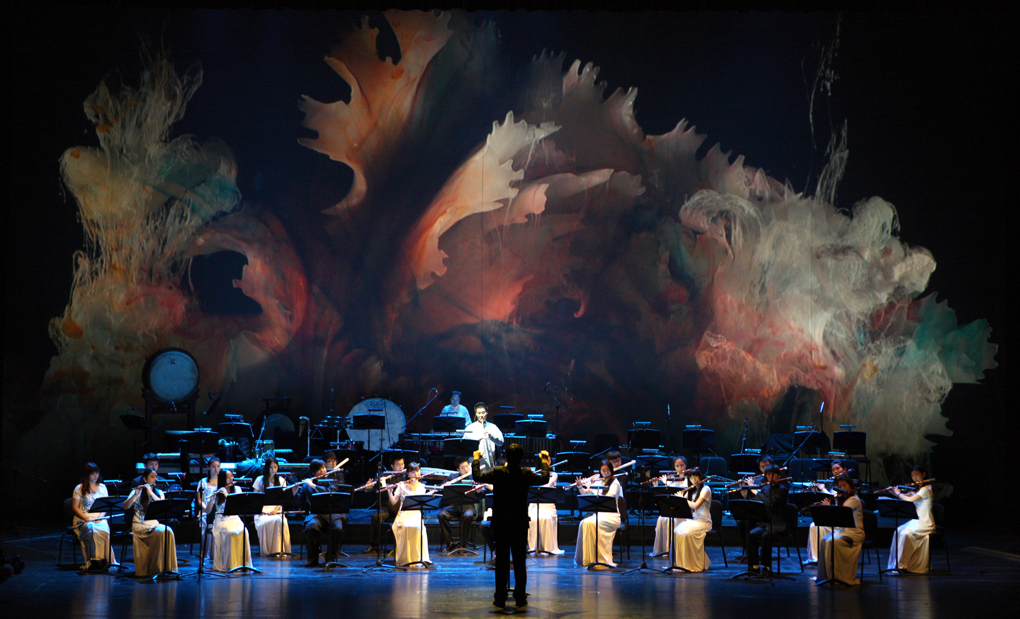 A large group of musicians playing Chinese bamboo flutes onstage with a conductor with a very colorful cloud-like image in the background.
