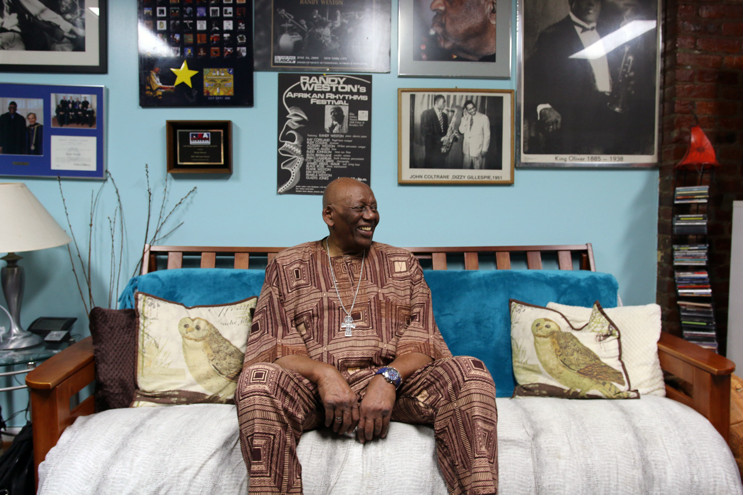Randy Weston sitting on his couch om front of many framed posters from performances, awards, etc.