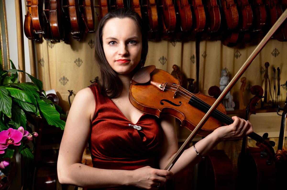Kinga Augustyn wearing a red dress to which a violin-shaped pin is affixed and holding a violin and bow in front of a shelf filled with violins.