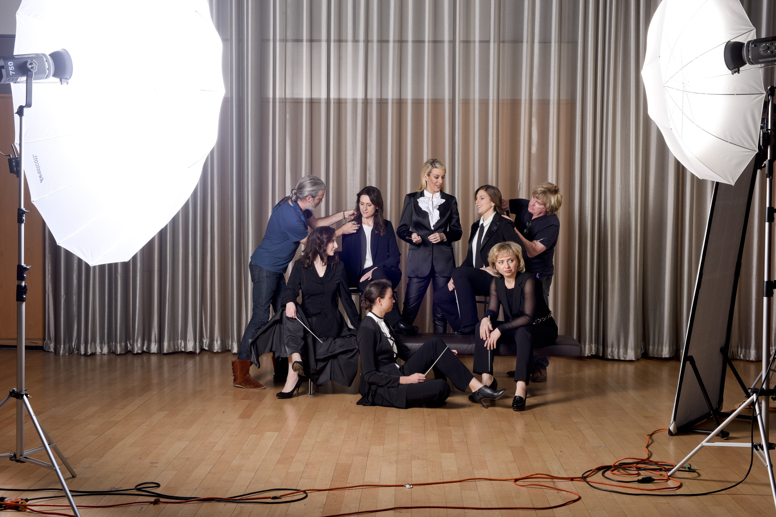 Six of the female conductors participating in the Hart Institute posing in tuxedos with batons for a photo shoot as two others help them adjust their positions for the camera.