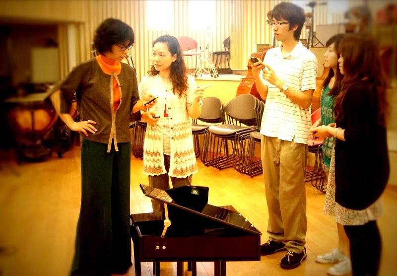 Ju-Ping Song and two students in a reheasal room standing around a toy piano positioned on the floor. (In the background are timpani and stacked chairs.)