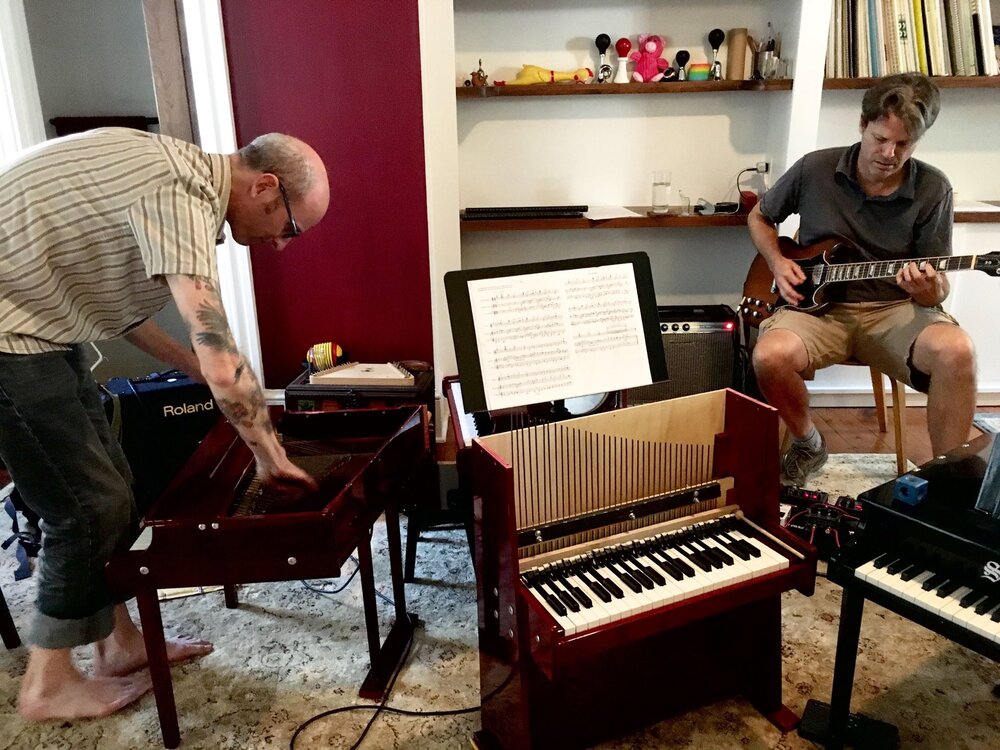 David Smooke leaning down toward a toy piano and Chad Kinsey sitting and playing electric guitar/