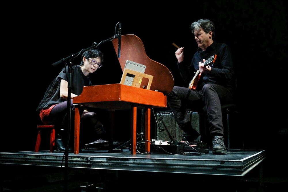 Ju-Ping Song sitting at a toy piano and Chad Kinsey standing playing electric guitar on a stage.