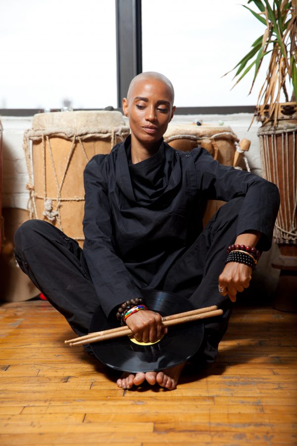 Val Jeanty holding drumsticks and sitting on a wooden floor near a window surrounded by large drums