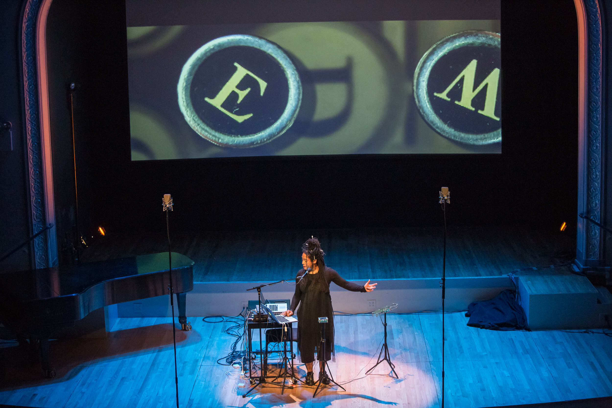 Pamela Z performing on two laptops with a microphone and various sensors standing on stage in front of a film projection of various letters in different directions.